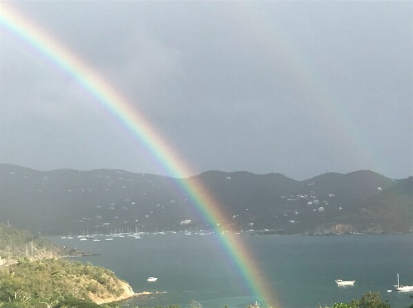Een kustlandschap met een regenboog boven een wateroppervlak, heuvels op de achtergrond en verschillende boten.
