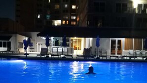 Une piscine entourée de bâtiments dont les fenêtres sont illuminées, avec des chaises longues et des parasols à proximité.