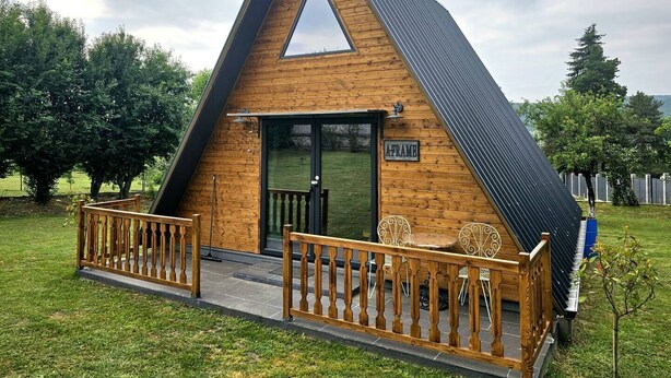Een houten cabin in driehoekige vorm met een glazen deur, een klein terras met stoelen en een bord waarop staat 