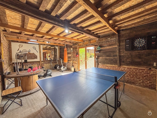 Une table de ping-pong dans une pièce rustique avec des poutres en bois, du matériel d’exercice et un jeu de fléchettes.