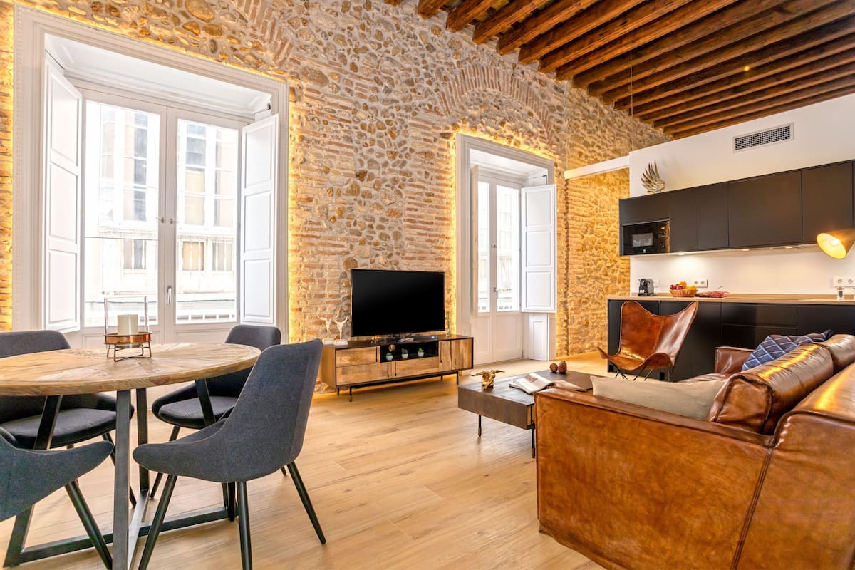 Un salon avec un mur en pierre, un plancher en bois, une table ronde avec des chaises, un canapé en cuir et un téléviseur à écran plat.