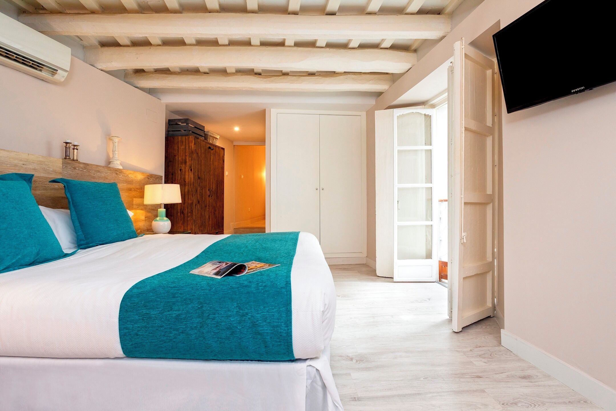 A hotel room with a bed, a TV, a wooden headboard, a lamp, and a door leading to another room.