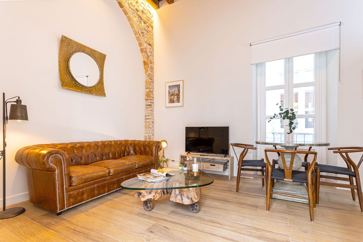 Un salon avec un canapé en cuir marron, une table basse en verre, un téléviseur et un coin repas avec des chaises en bois.