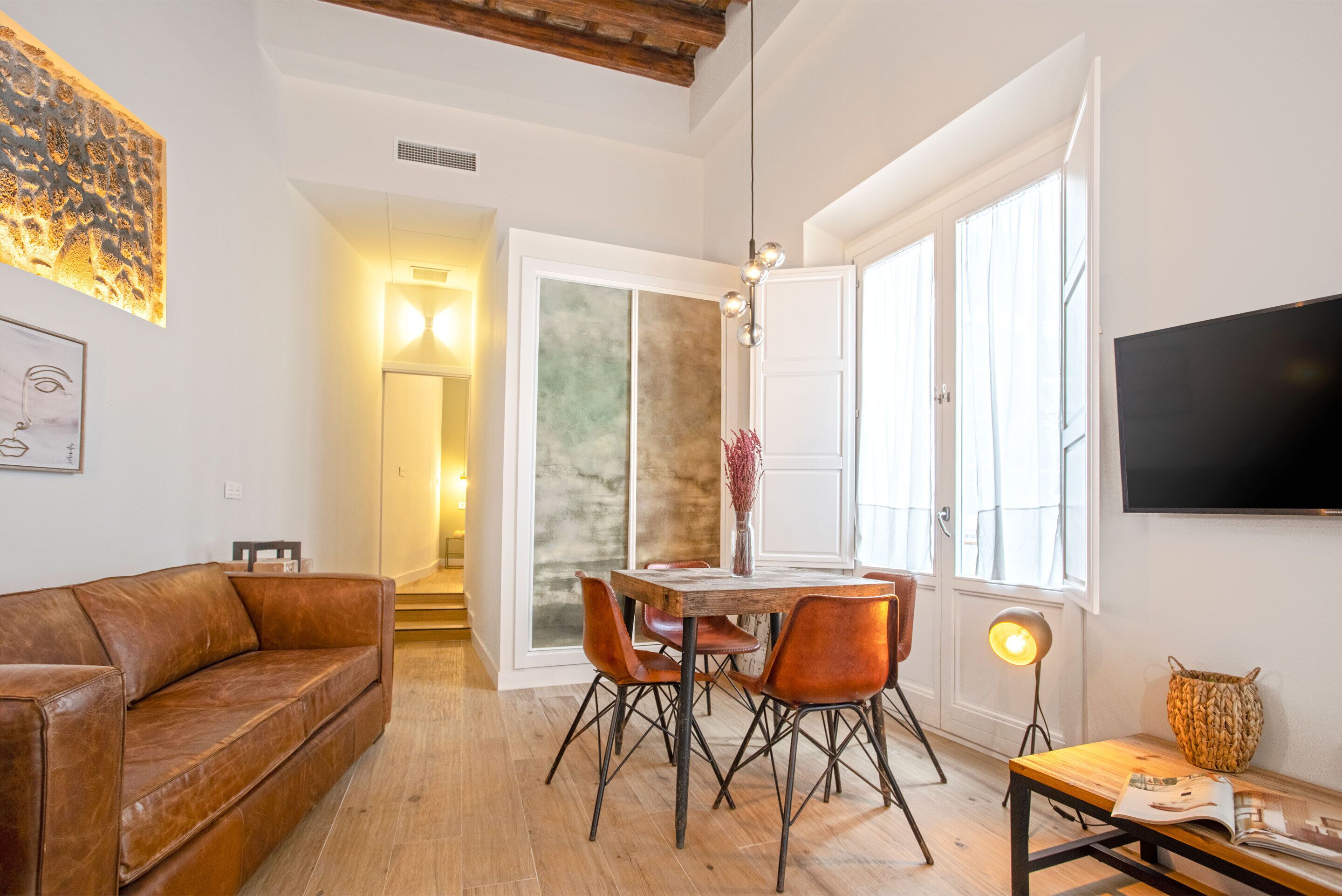 Ein modernes Wohnzimmer mit einer braunen Ledersofa, einem Holzesstisch mit vier Stühlen, einem flachbildfernseher an der Wand und einem großen Fenster mit Vorhängen.