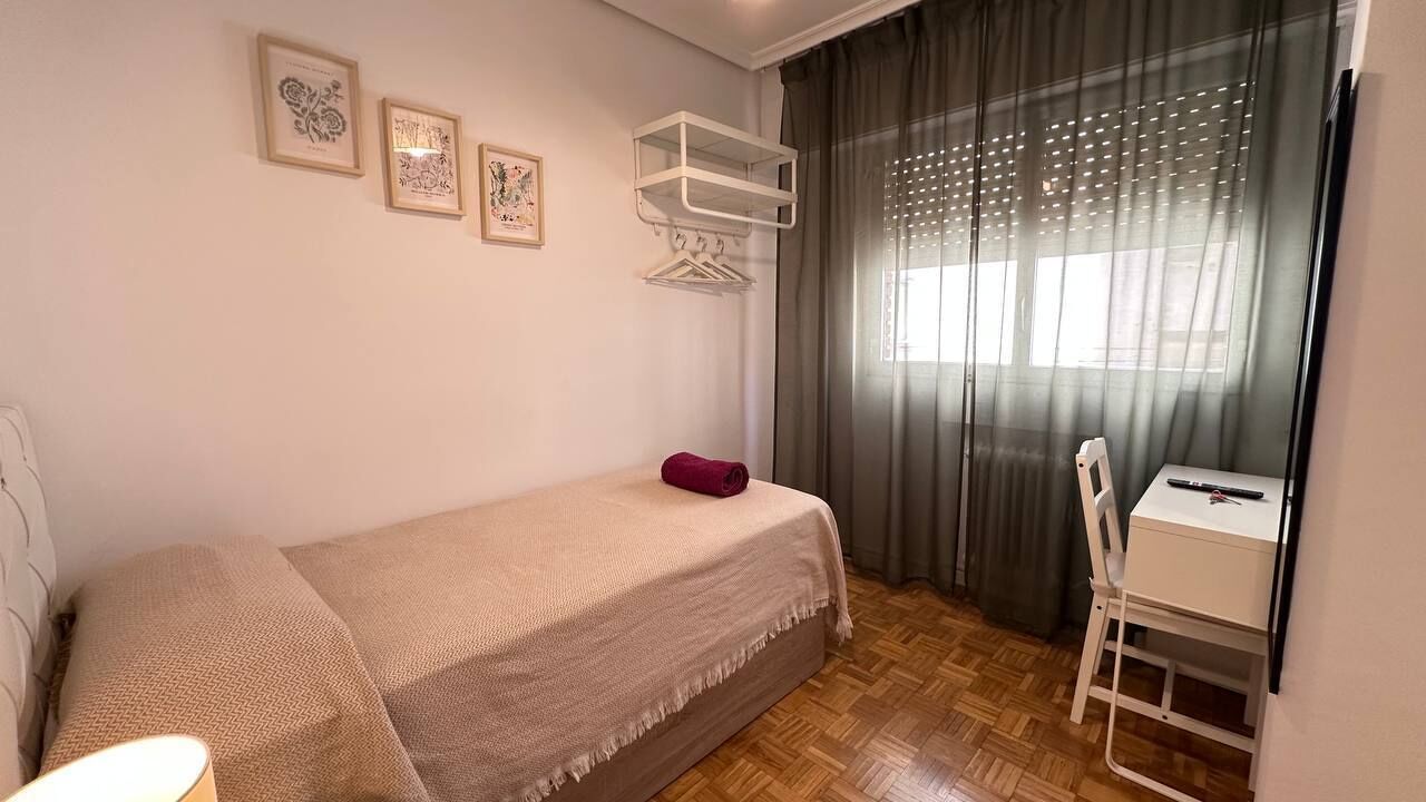 A small, neatly arranged room with a bed, a desk, and a chair. There are two framed pictures on the wall, a shelf above the bed, and a window with curtains.
