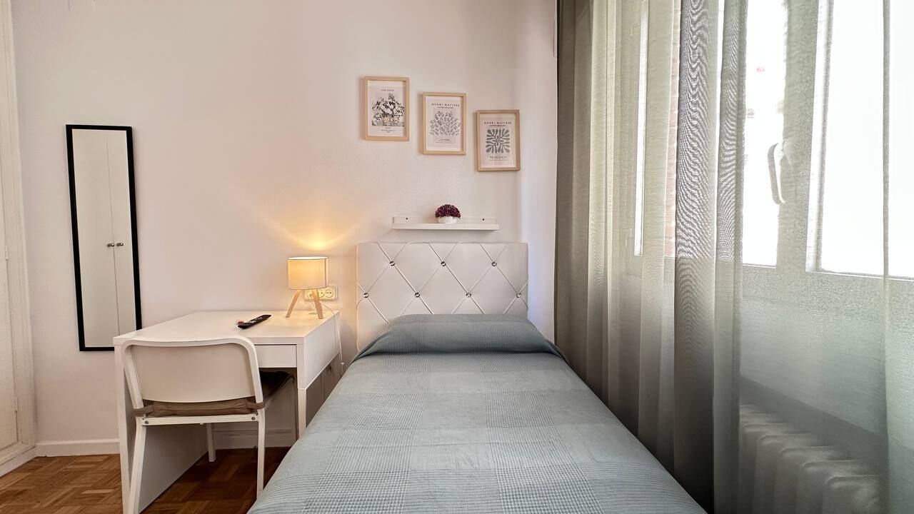A bedroom with a bed, a desk, a chair, a bedside table, a mirror, and a window with curtains.