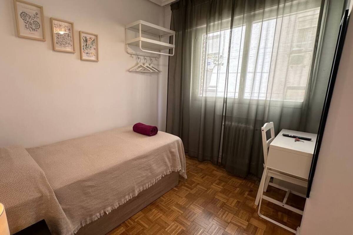 Ein kleines, ordentlich eingerichtetes Schlafzimmer mit Bett, Nachttisch, Stuhl und einem Fenster mit Vorhängen.