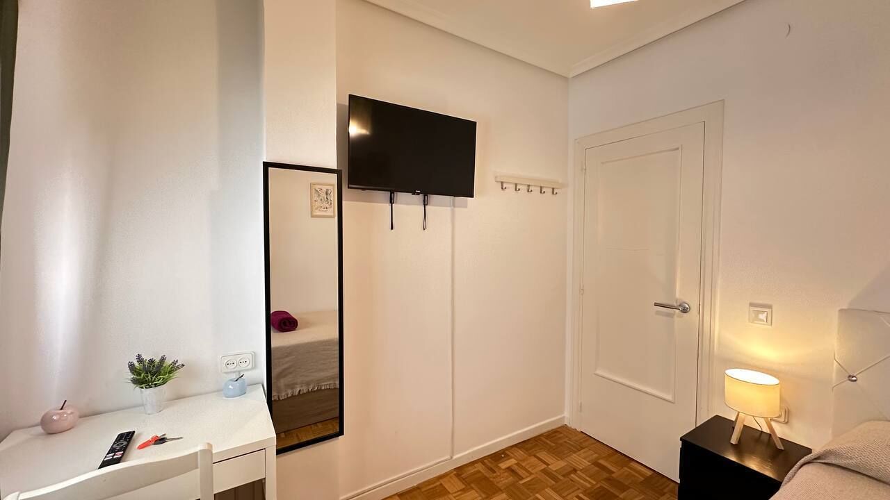 A bedroom with a bed, bedside table, lamp, TV, and a mirror.