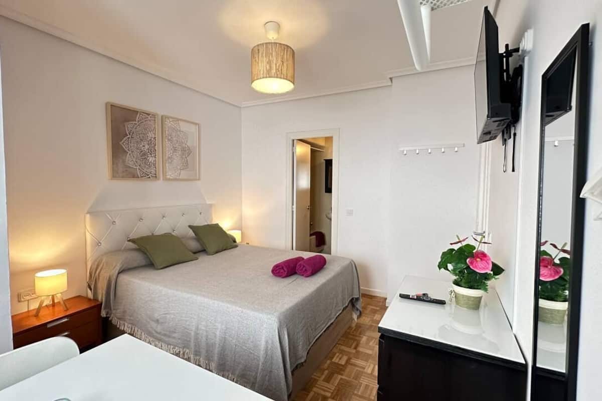 Ein Schlafzimmer mit Bett, Nachttisch, Lampe und einem an der Wand befestigten Fernseher.