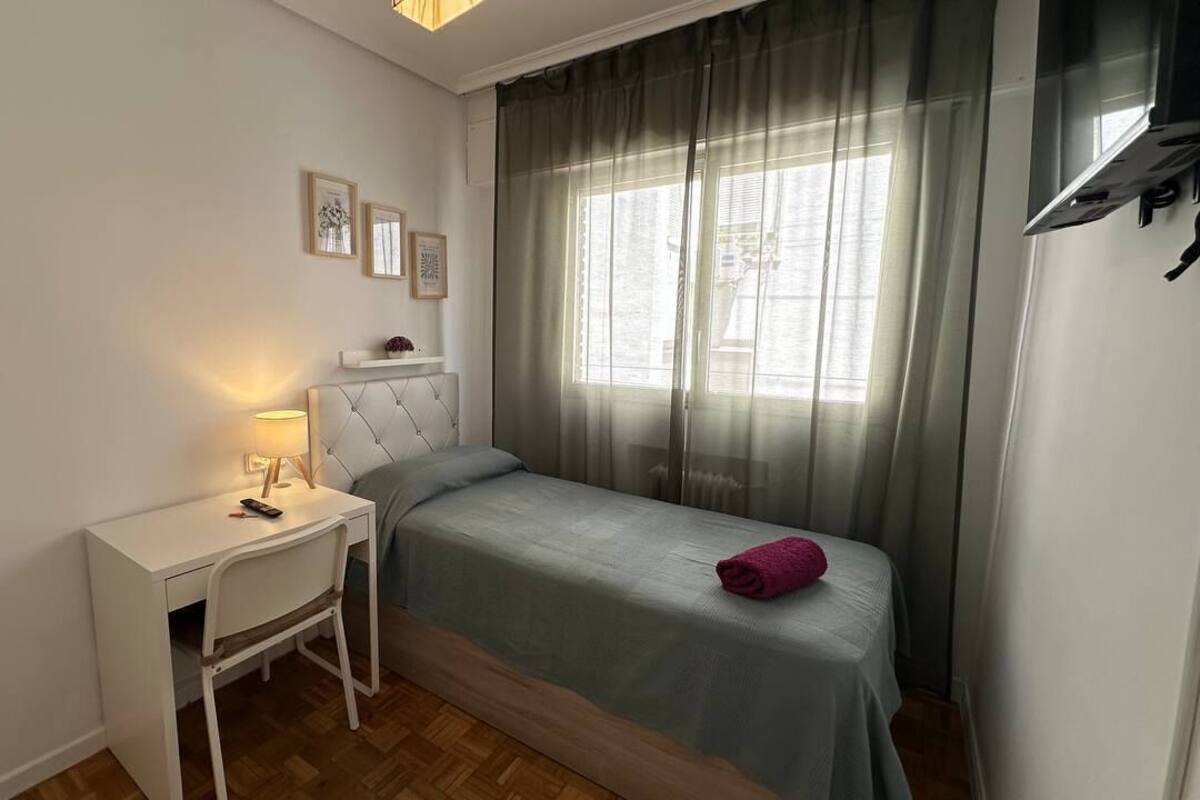Ein Schlafzimmer mit einem Bett, einem Schreibtisch, einem Stuhl, einem Fenster mit Vorhängen und Wandbildern.