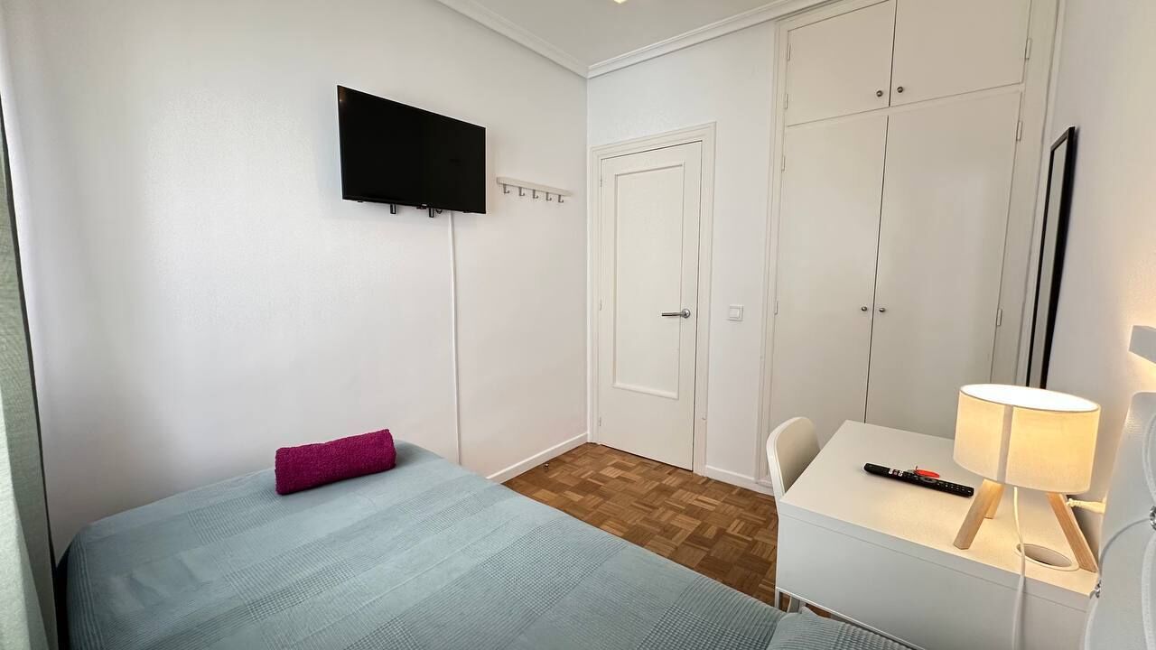 Een kamer met een bed, een bureau, een stoel, een aan de muur bevestigde televisie en een garderobe met spiegel.