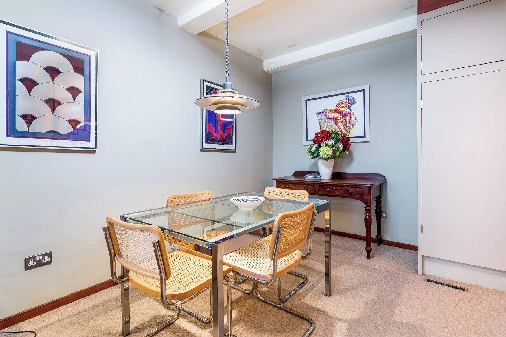 A dining area with a glass table, wicker chairs, a wooden side table with a vase of flowers, and artwork on the walls.