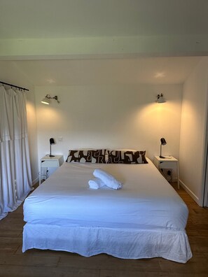 Una cama grande con ropa de cama blanca, dos lámparas de noche, un cojín decorativo y una toalla doblada sobre la cama.