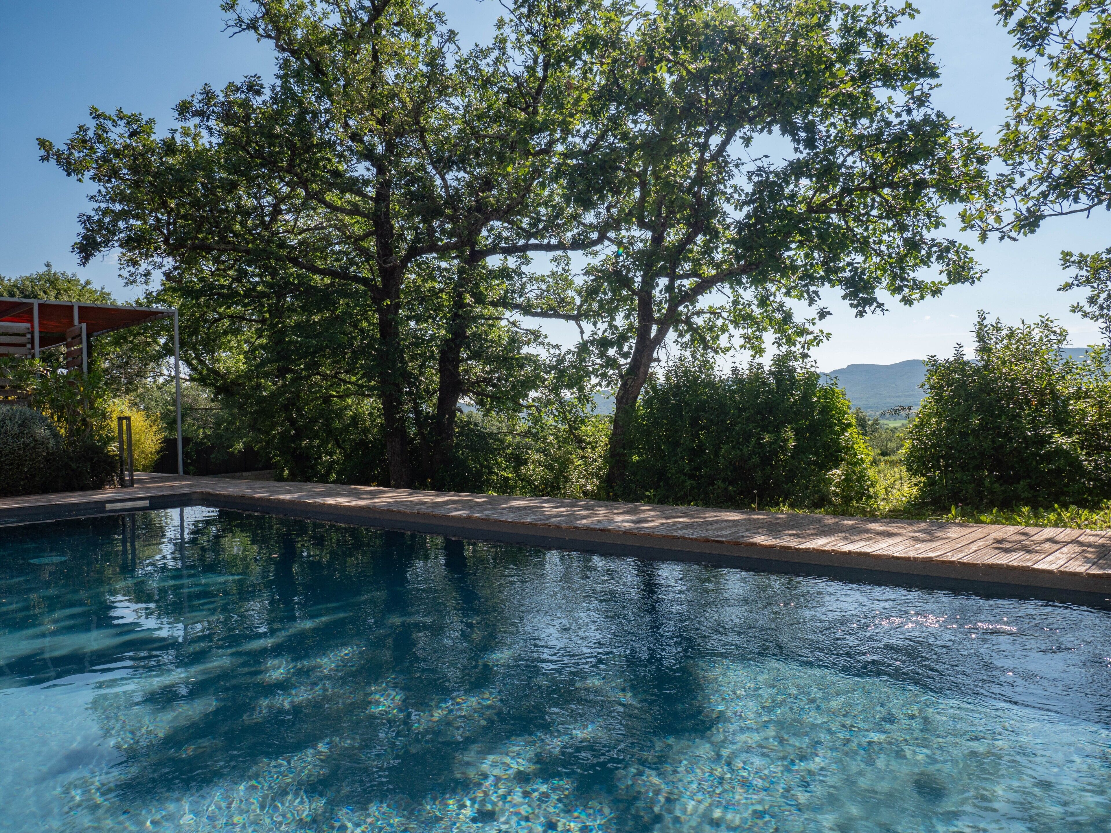 A swimming pool with clear water, surrounded by trees and a view of distant hills.