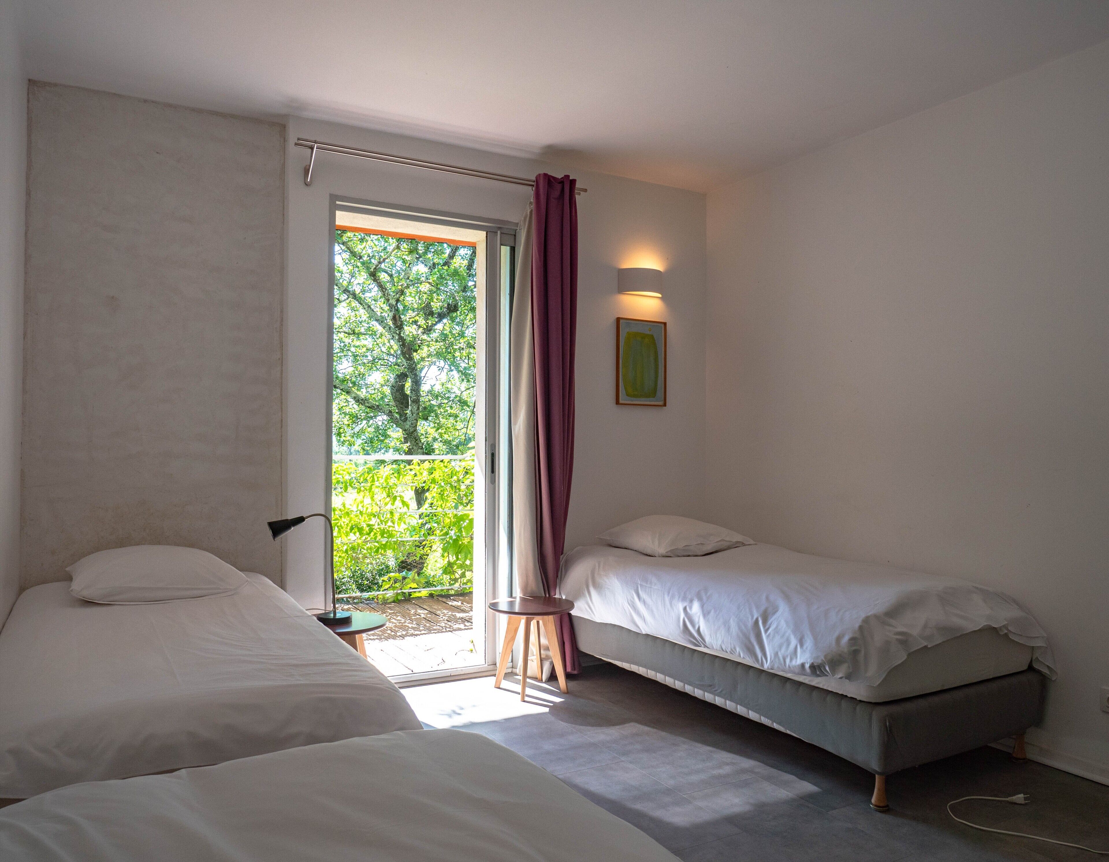 A bedroom with two beds, a window with curtains, a small table, and a wall-mounted light fixture.
