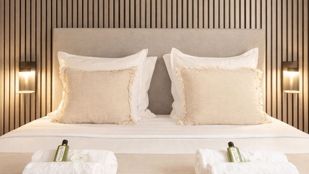 Camera d'albergo con un letto grande, due cuscini, due luci a parete e due asciugamani con bottiglie sul letto.