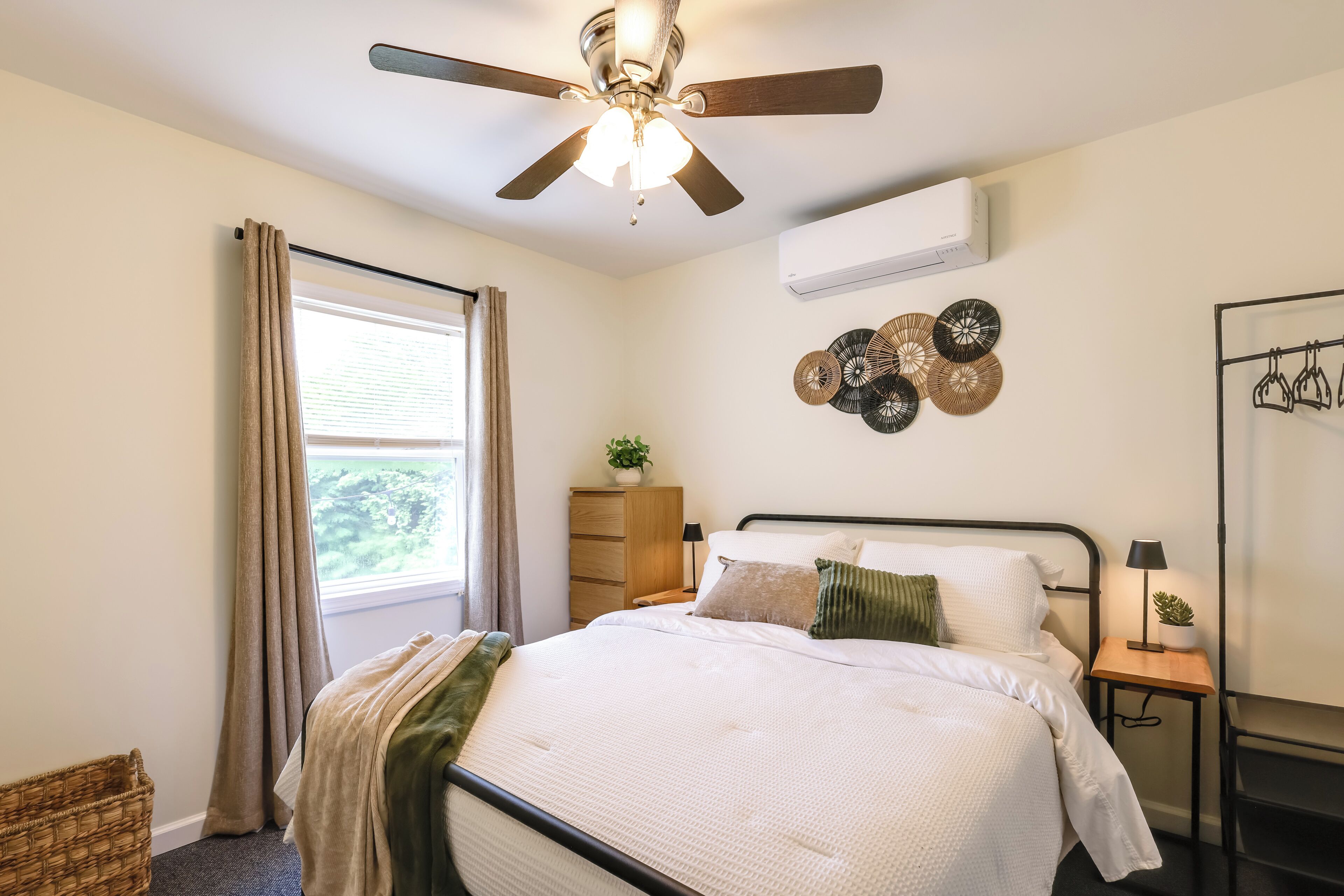 Une chambre à coucher comprenant un lit, un ventilateur de plafond, une fenêtre avec des rideaux, une table de chevet avec une lampe et des éléments de décoration murale.