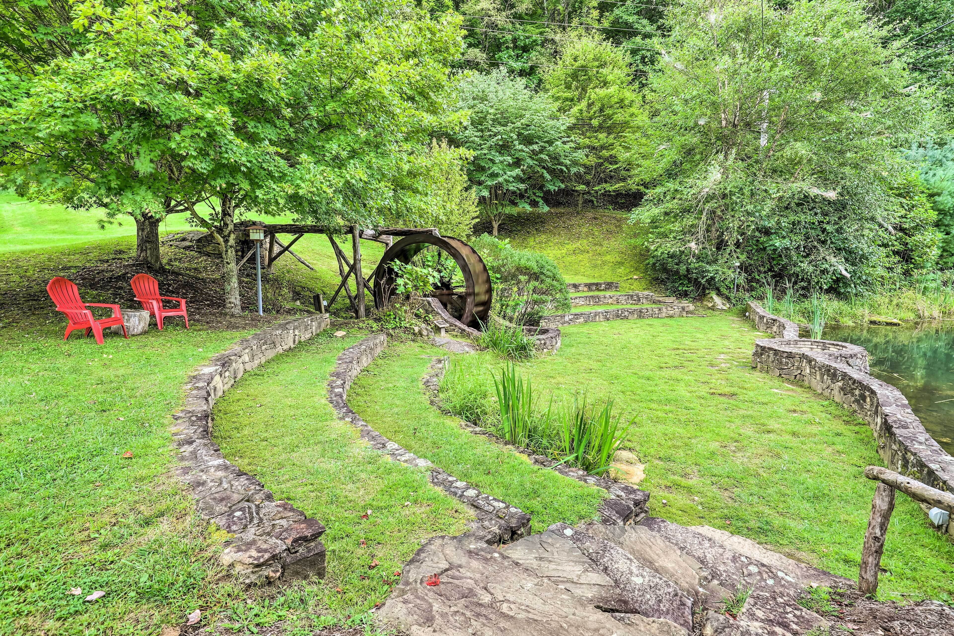 Un jardín con una rueda hidráulica, senderos de piedra y sillas rojas.