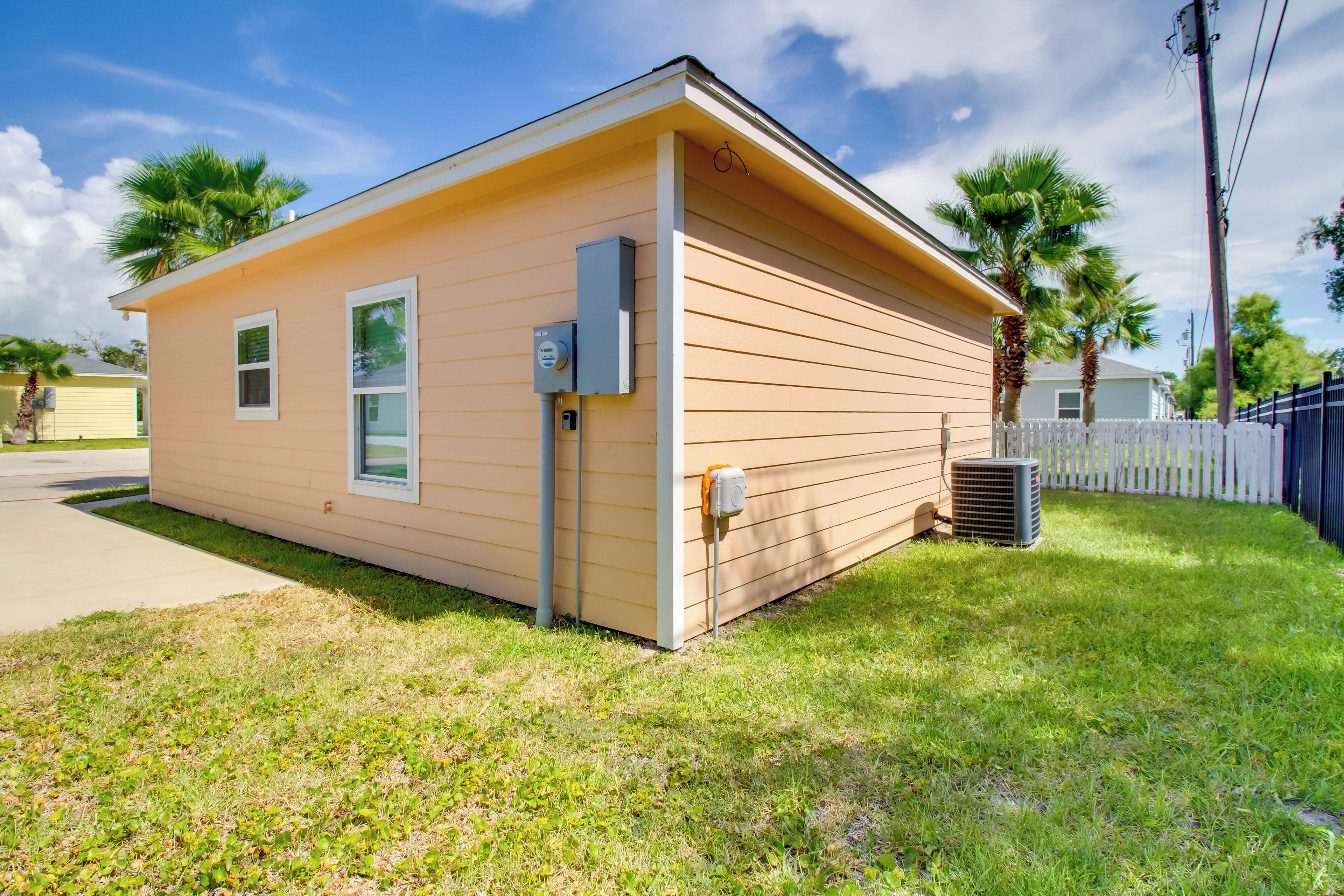 A single-story house with a small porch, a utility box, and a white picket fence.