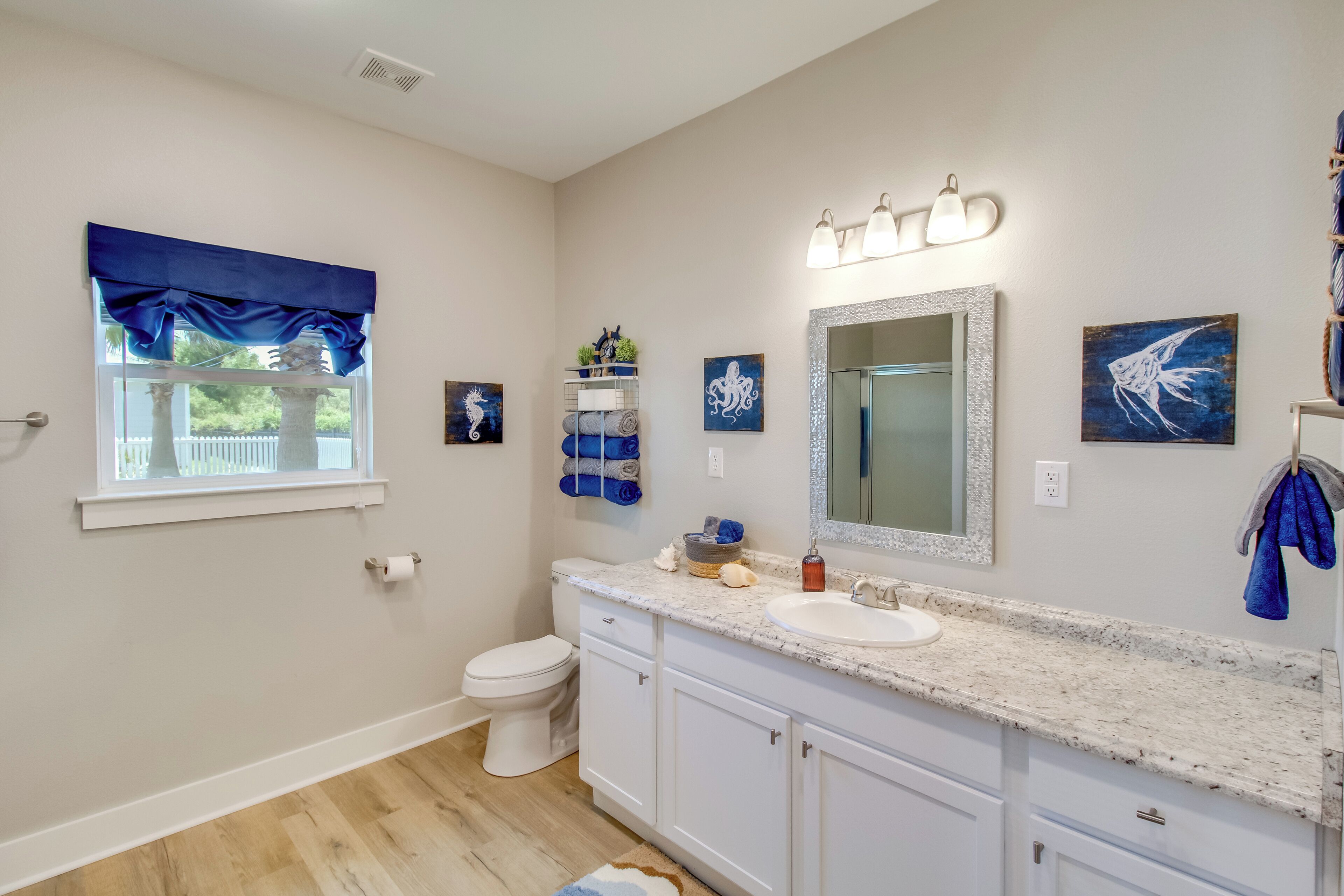 A bathroom with a toilet, sink, and vanity. There is a window with blue valance, a mirror with lights, and artwork on the walls.