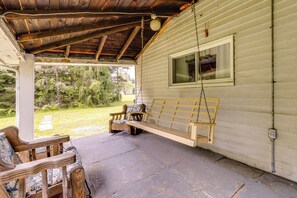 A covered porch with a wooden bench and chairs, a hanging swing, and a view of a forested area. - Pet-friendly Smithmill Retreat w/ Yard! (Houtzdale)