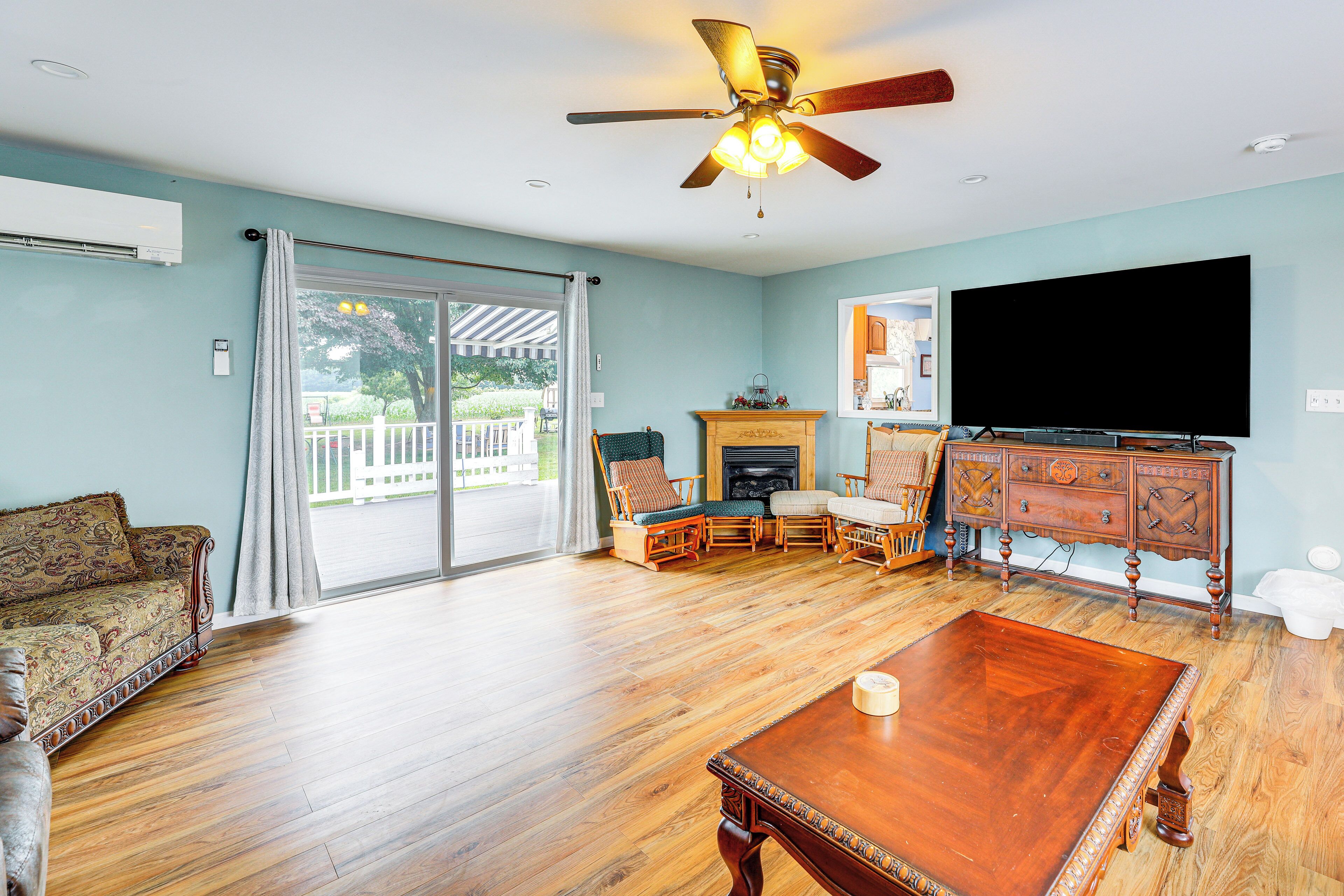 A spacious living room with a wooden floor, a ceiling fan, a flat-screen TV, a fireplace, and a dining table.