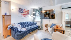 Un salón con un sofá azul, un sillón blanco, un aparador de madera, un televisor y una ventana con cortinas.