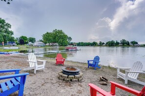 A lakeside setting with several Adirondack chairs, a fire pit, and a dock with boats. - Peaceful Retreat on Gilead Lake w/ Grill! (Bronson)