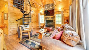 L’intérieur d’une cabane en rondins, avec un escalier en colimaçon, une cheminée en pierre, des meubles en bois et un coin salon confortable.