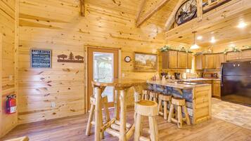Interior de una cabaña de madera con cocina, taburetes de madera, extintor y un reloj en la pared.