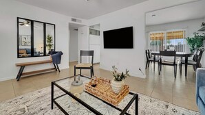 Una sala moderna con ventilador de techo, televisor de pantalla plana, comedor con mesa y sillas, y un banco con planta.