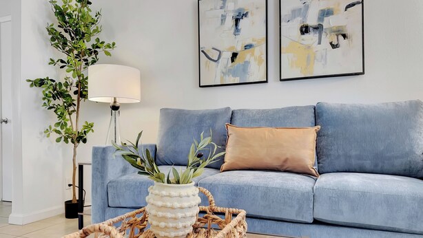 Sala de estar con un sofá azul, una lámpara blanca, dos cuadros abstractos y una planta en maceta sobre una bandeja tejida.