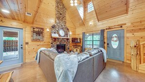 A log cabin interior with a stone fireplace, a large sofa, and a wooden ceiling. - Near Choctaw Landing 2 King Bedrooms Game Loft (Broken Bow)