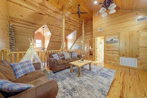 A cozy log cabin interior with a loft bedroom, a living area with a sofa and coffee table, and a stone wall feature. - Near Choctaw Landing 2 King Bedrooms Game Loft (Broken Bow)