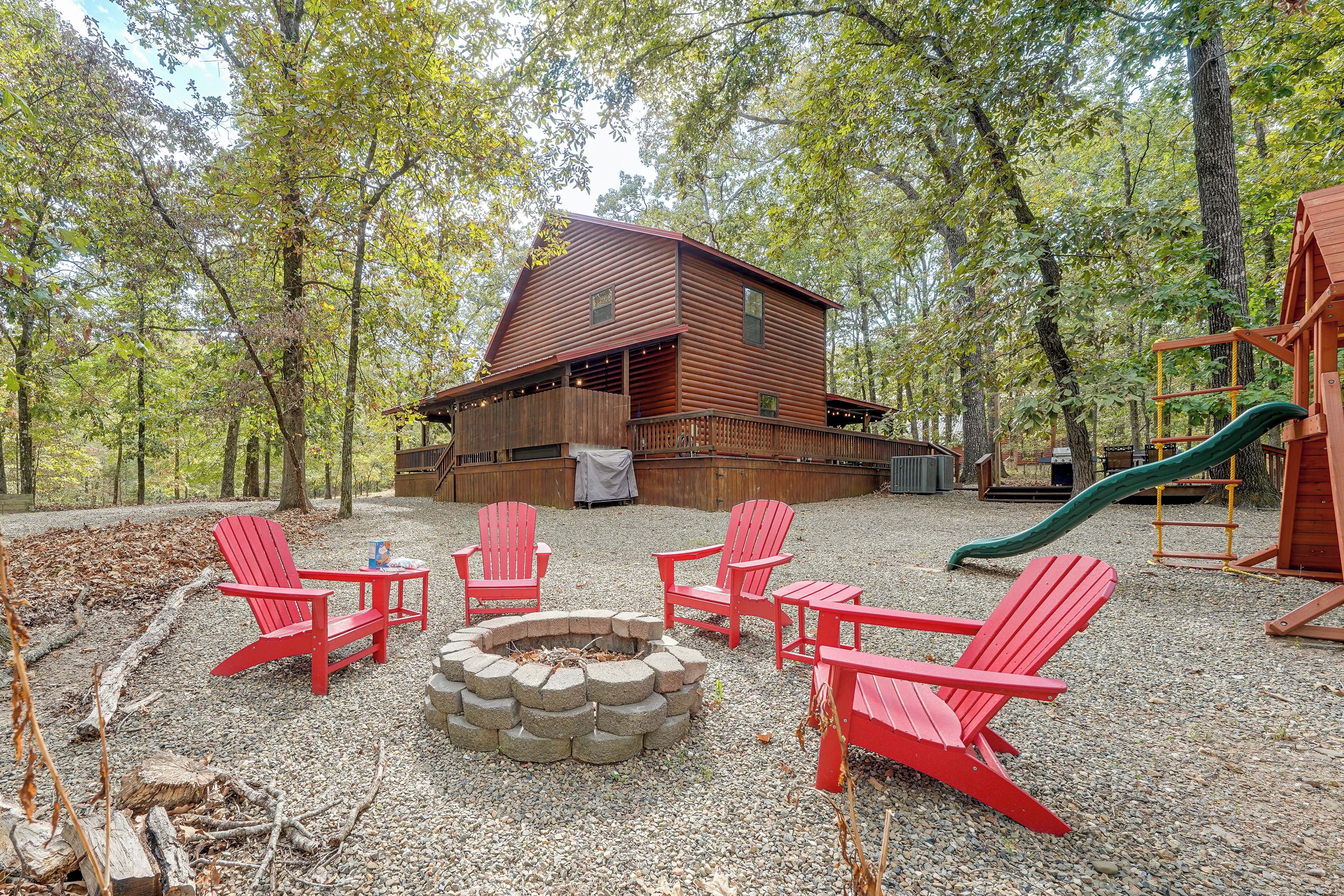 A cabin with a deck, surrounded by trees, and a fire pit with red Adirondack chairs.