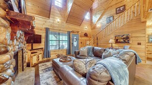 A log cabin interior with a stone fireplace, leather sofa, wooden walls, and a staircase. - Near Choctaw Landing 2 King Bedrooms Game Loft (Broken Bow)