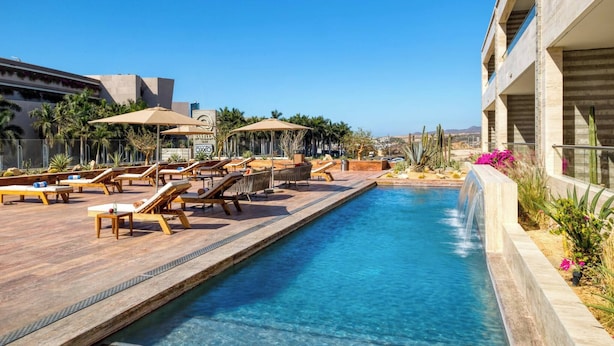 A pool area with lounge chairs and umbrellas, surrounded by palm trees and a building with a modern design.