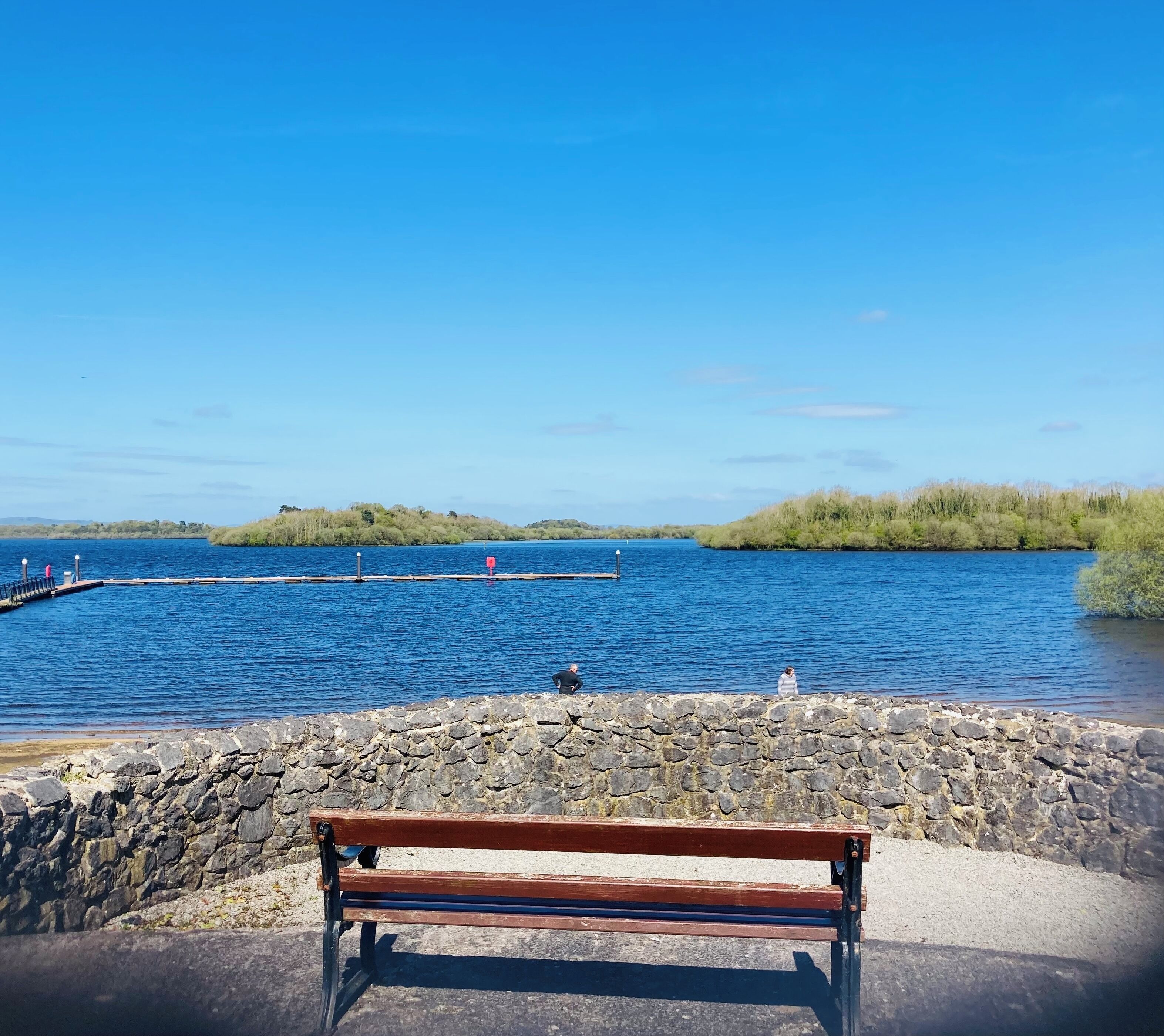 Un banc en bois, adossé à un mur de pierres, surplombe une étendue d’eau calme, et une jetée s’avance sur cette eau.
