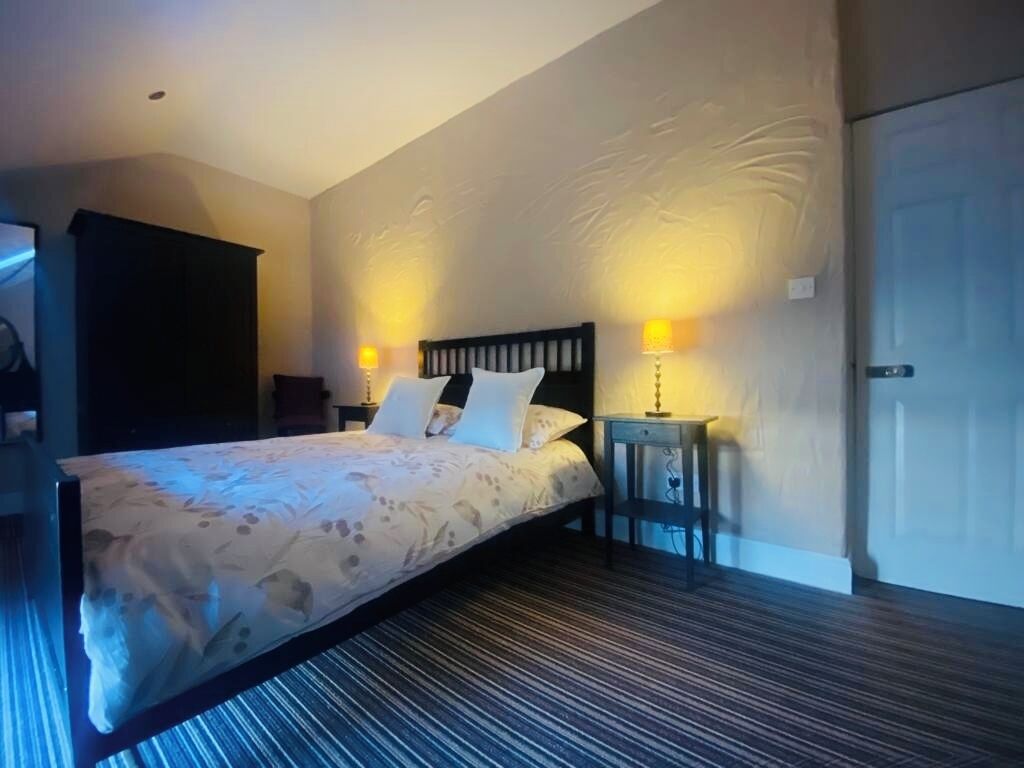 A hotel room with a bed, two bedside lamps, a dark wooden headboard, a small bedside table, and a door.
