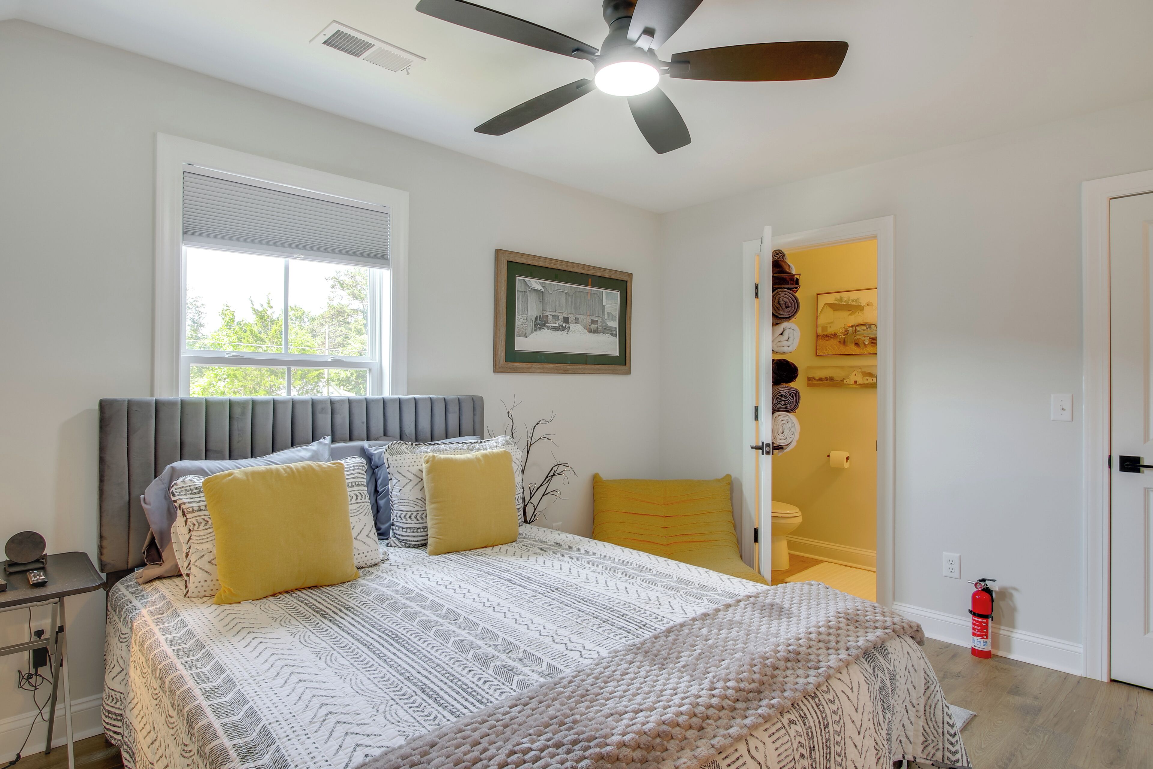 Un dormitorio con cama, ventilador de techo, ventana, un cuadro enmarco y una puerta que da a un baño.