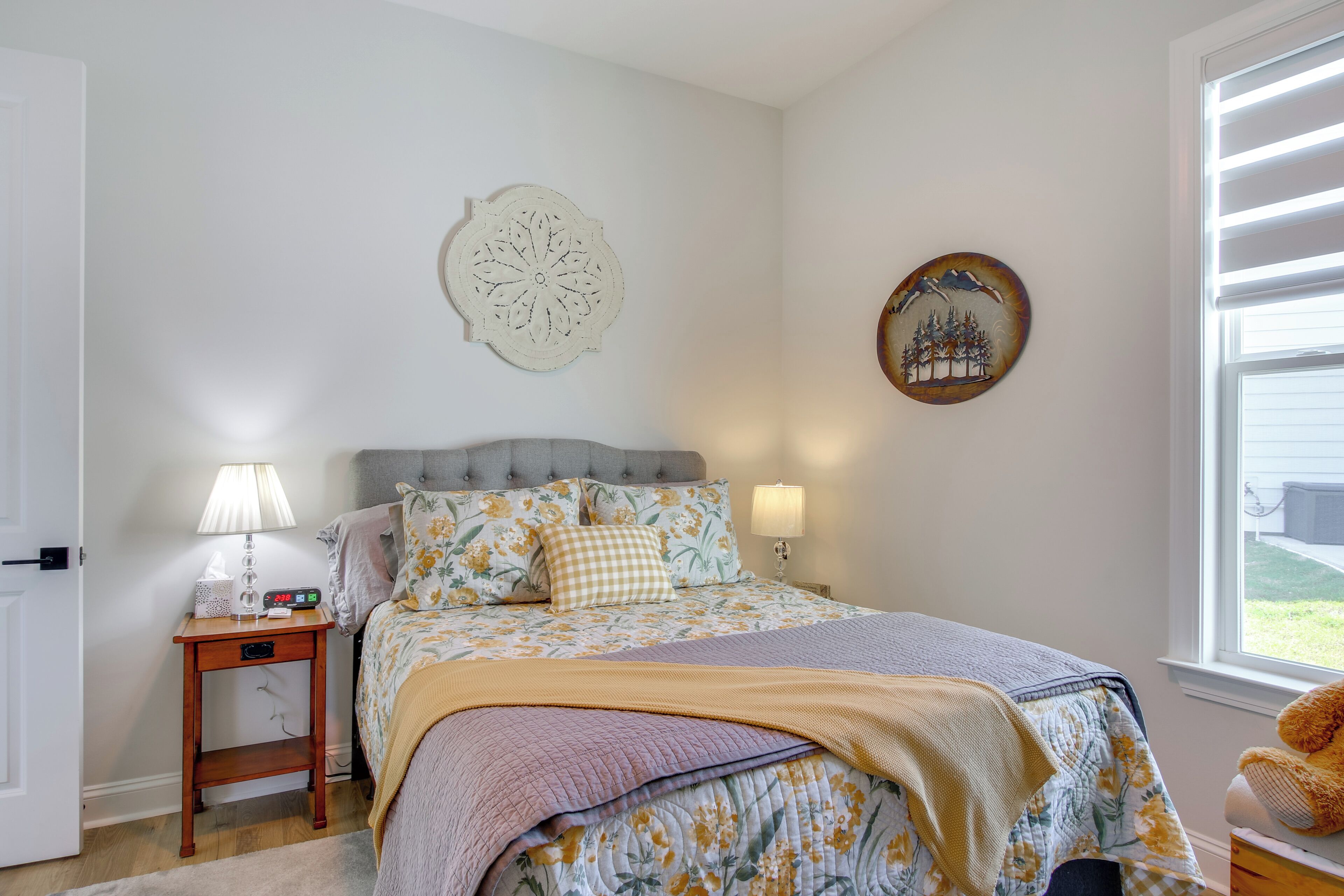 Quarto com cama, abajures, mesa de cabeceira, um objeto decorativo na parede e uma janela com persianas.