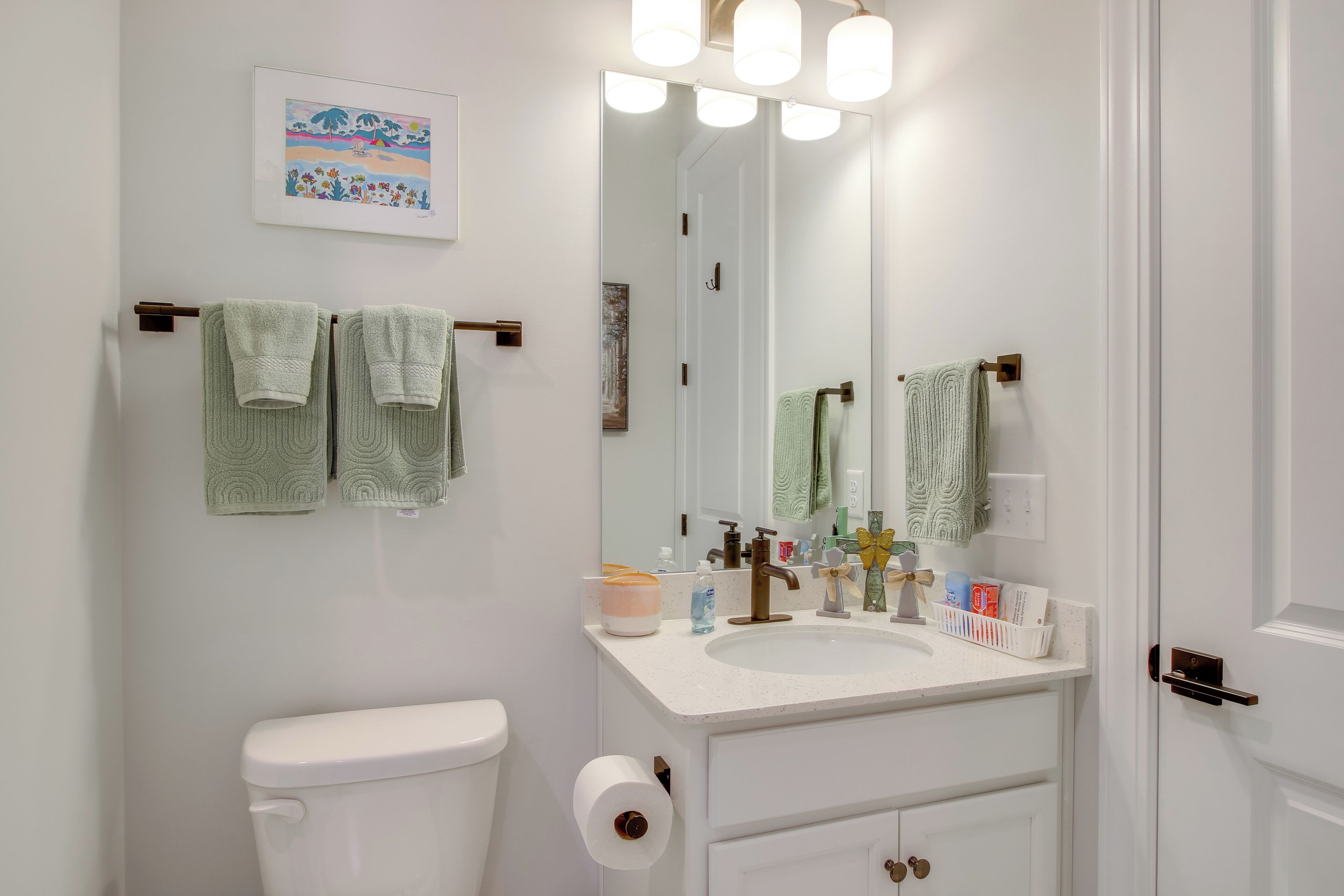 Un baño con inodoro, lavabo y espejo. Hay toallas colgadas en un perchero y artículos de aseo en la encimera del lavabo.