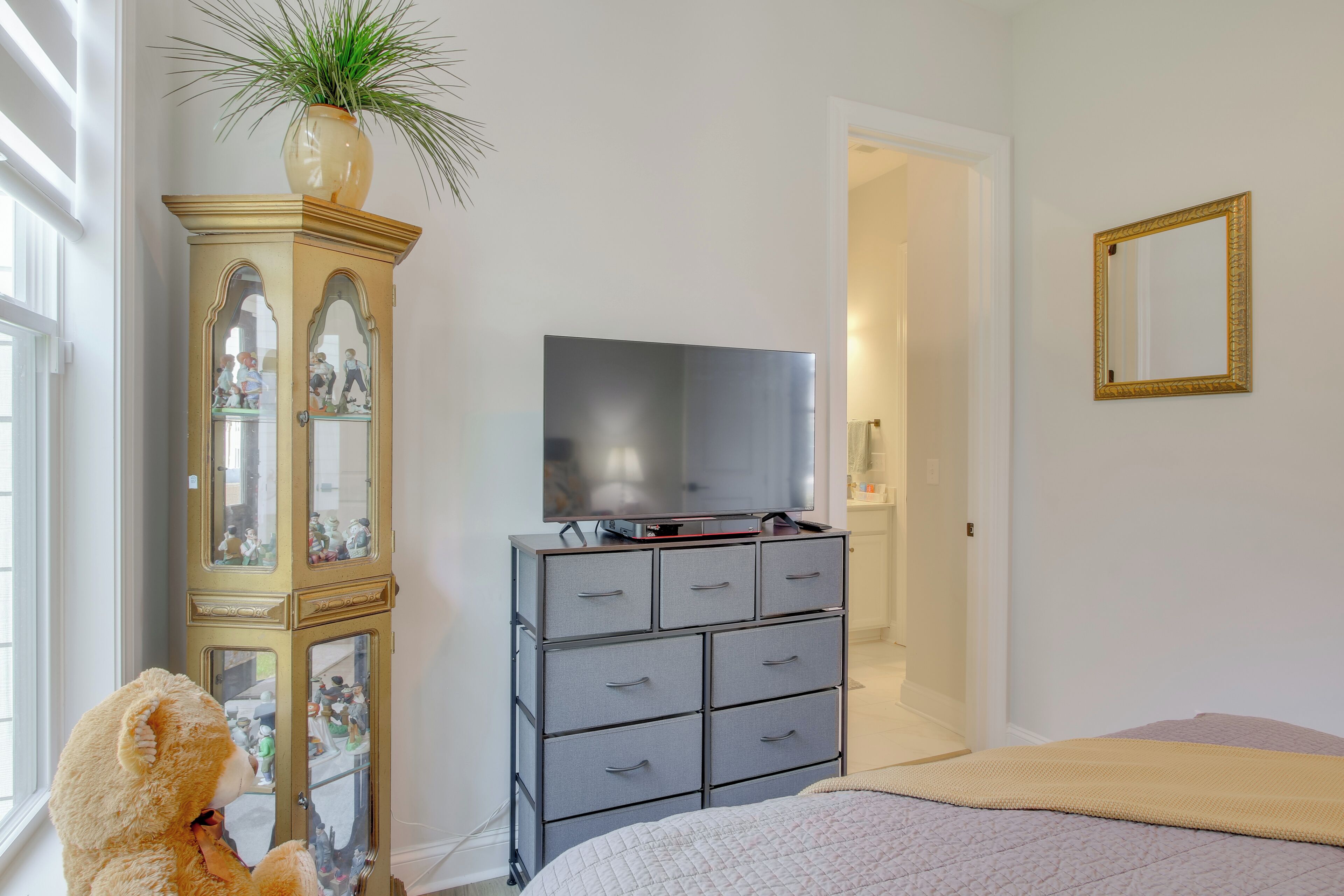 Un dormitorio con cama, cómoda con televisor, espejo y ventana con persianas.