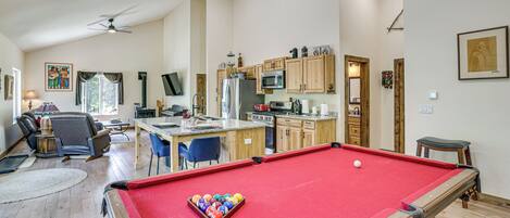 A spacious living area with a pool table, a kitchen with wooden cabinets, and a dining area with blue chairs.