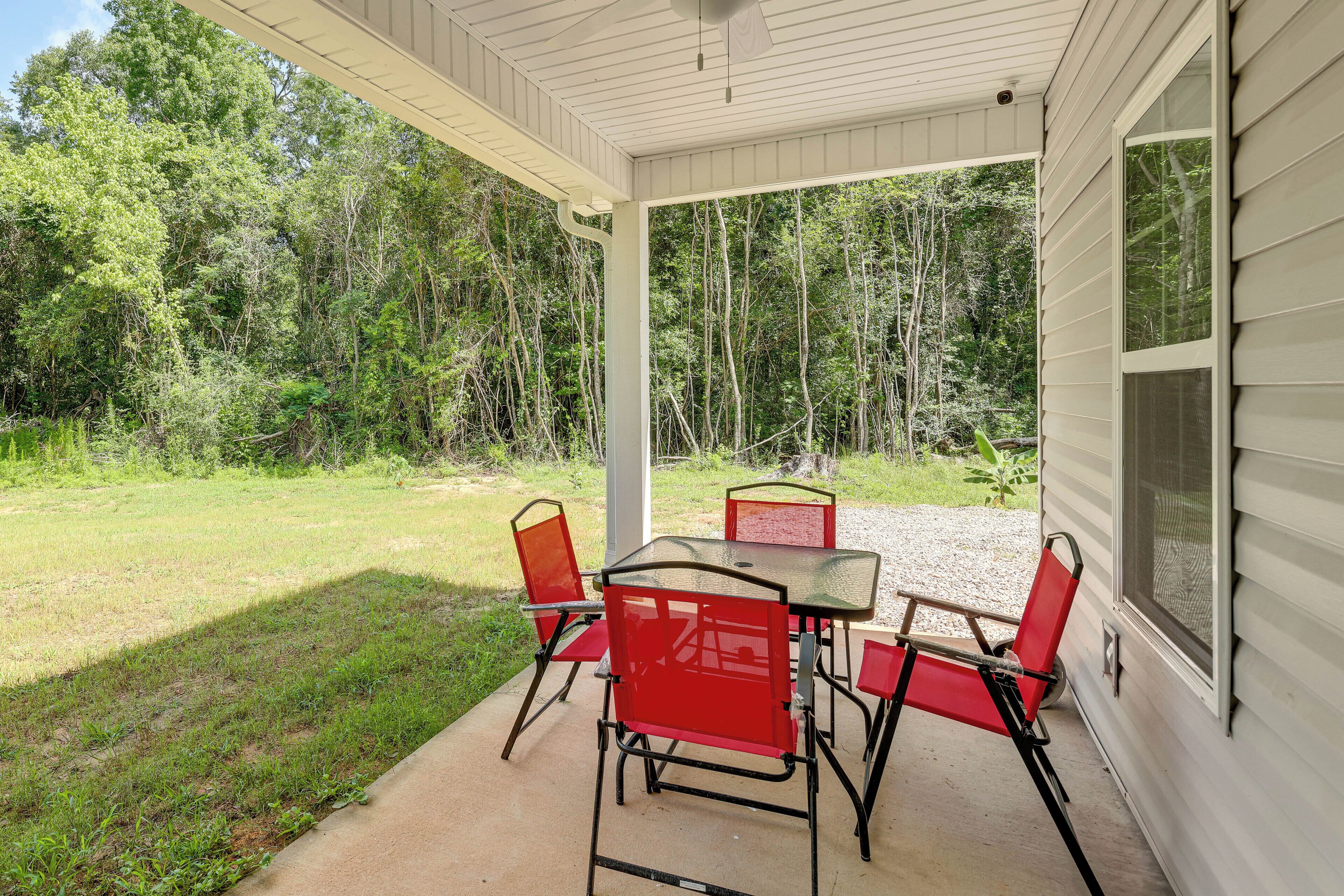 A covered patio with a table and four red chairs, surrounded by a grassy area and trees.
