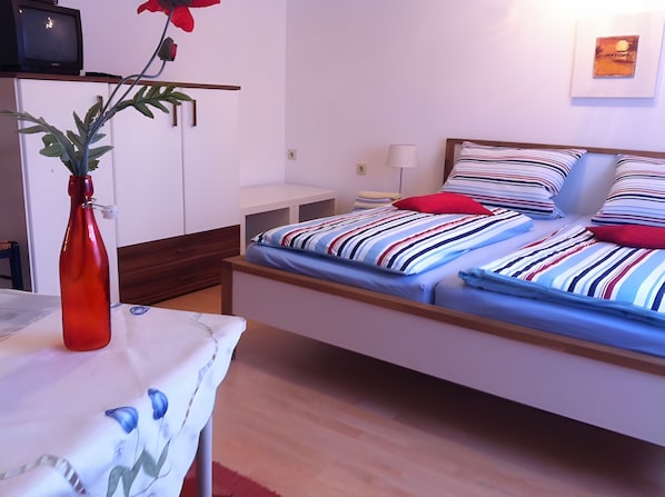 A bedroom with a bed, a nightstand, a lamp, a TV, and a red vase with a plant.