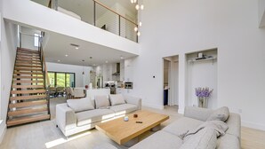 A spacious living area with a high ceiling, a wooden staircase, a large sofa, a coffee table, and a kitchen in the background. - Walk to Celebration Park: Naples Home w/ Pool! (Naples)