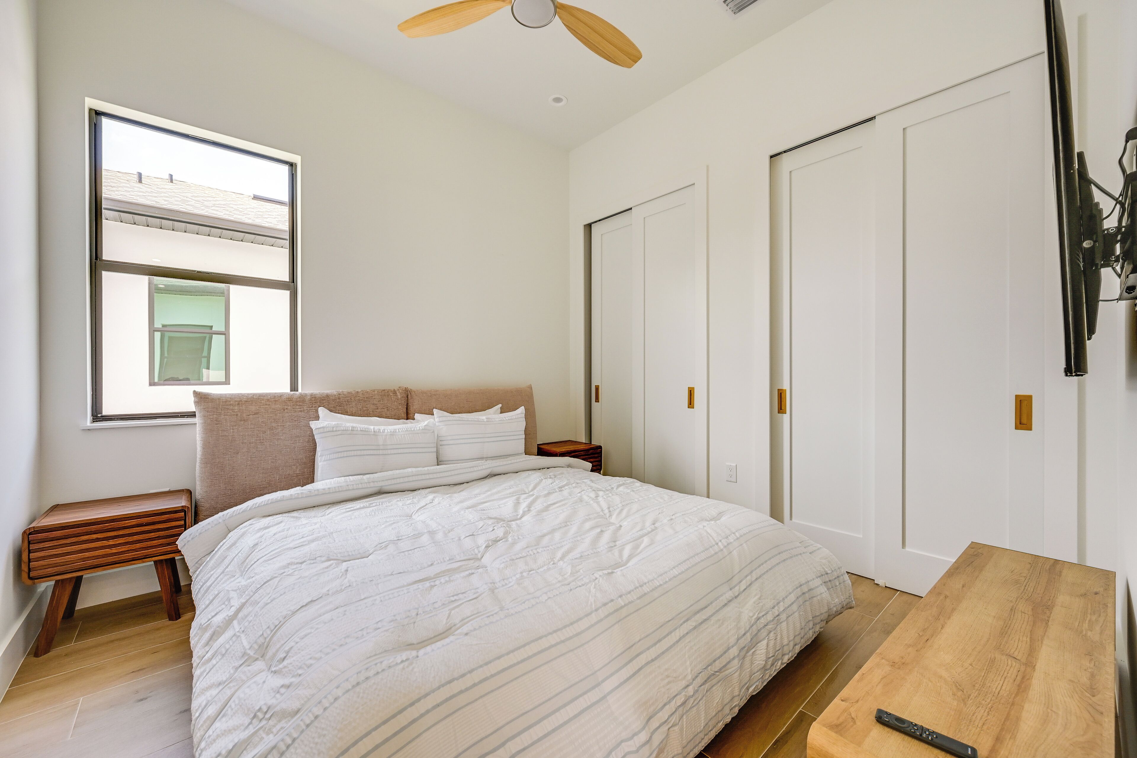 Une chambre à coucher comprenant un lit, une table de chevet, une table en bois, un ventilateur de plafond et une fenêtre.