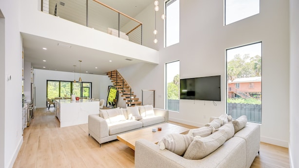 Een ruime woonkamer met een hoge plafond, een trap naar een hoger gelegen verdieping, een grote flatscreen televisie aan de muur en uitzicht op de omgeving door grote ramen.