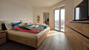 Un dormitorio moderno con una cama de madera, un televisor sobre un mueble de madera y un balcón con vista a la ciudad.